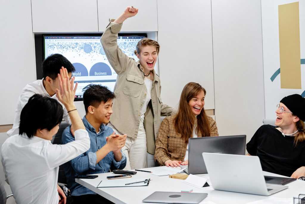 A diverse team enthusiastically celebrating success around laptops during an office meeting.