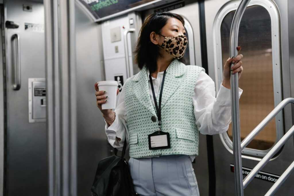 Asian woman wearing a facemask holding coffee on a subway train, depicting the new normal of commuting.